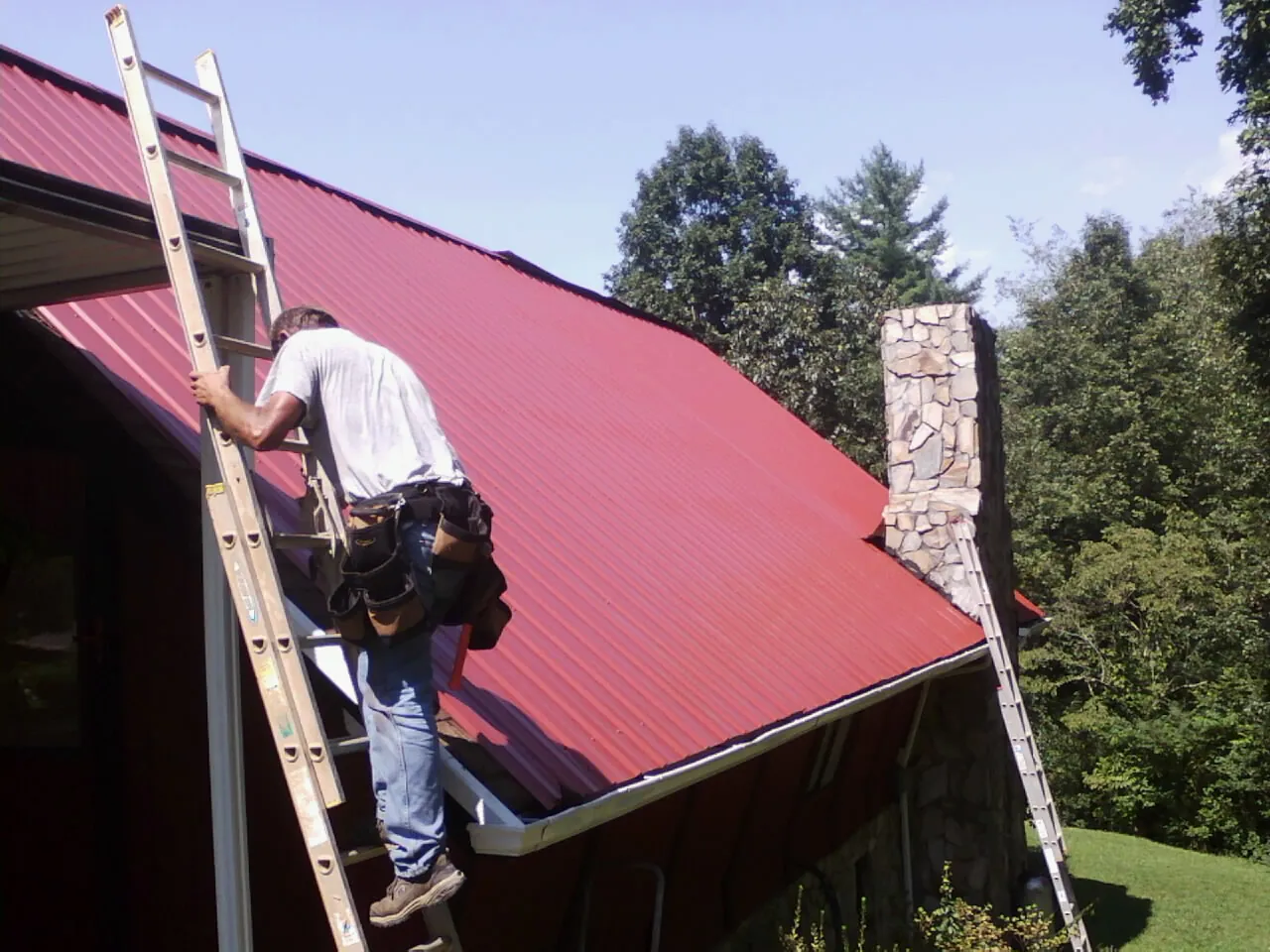 Roofer on a ladder beside a red metal roof and stone chimney — Soffit & Fascia Repair by Roofing Brothers in Hazelwood